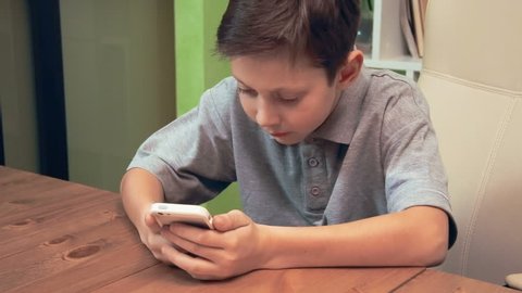 Little boy sends text on the phone sitting in the office chair at the table