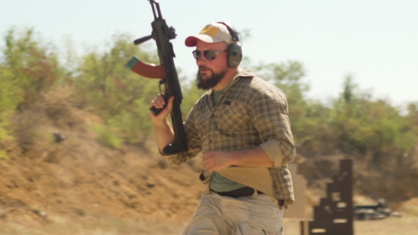 Man in headphones and with gun running behind car while another male aiming firing shotgun outdoors in slowmotion