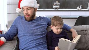 Father in christmas hat sitting on sofa near the son which reading book and dresses christmas hat on him