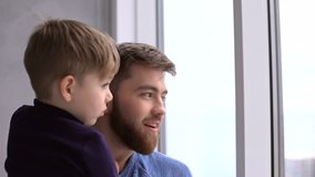 Smiling young father standing by window holding his little son and pointing finger at home 