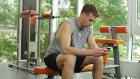 Muscular young man using mobile phone while sitting on the bench after the training in the gym