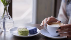 Young smiling woman enjoying coffee and tasty macaroon while sitting in cafe