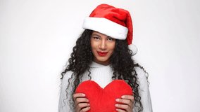 Shy African woman in white sweater and christmas hat holding plush heart in hands in studio