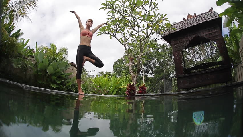 Split underwater shoot of young woman stretches and performs yoga exercises in the green graden by the pool