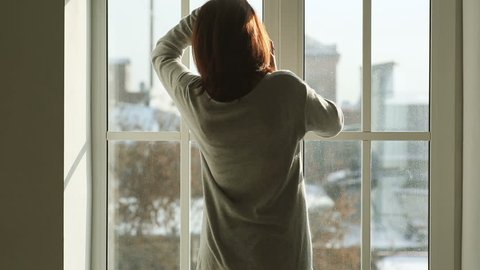Rear view of young woman in gray cardigan using mobile phone while standing near big window at home. In the beginning she talking on the phone and then ends call and starts typing message.