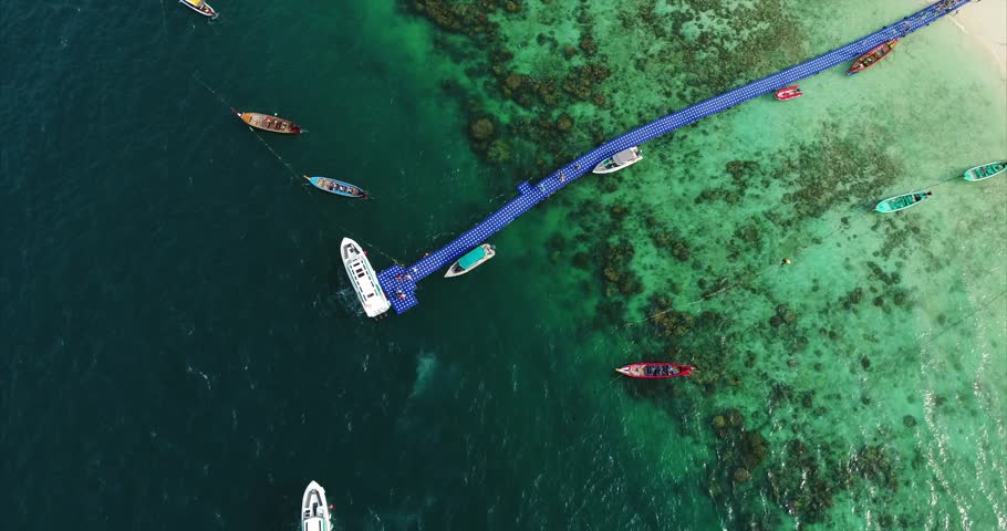 Top-down aerial view of passengers leaving boats at a bright floating dock in tropical water.