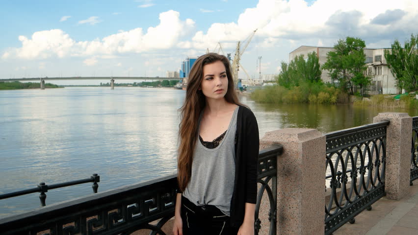 Urban waterfront sophistication: contemplative young woman in twenties stands on scenic river promenade, gazing thoughtfully toward expansive water with serene composure. She wears layered ensemble featuring soft gray tank beneath structured ebony cardigan, creating subtle contrast and visual depth. Her posture leans against ornate wrought-iron railing with geometric lattice pattern, suggesting European architectural heritage while long chestnut hair catches natural illumination. Setting showcases industrial waterfront landscape with calm river reflecting clouds, construction cranes and port infrastructure indicating active maritime commerce. Historic stone embankment architecture frames scene with cream-toned construction, modern residential structures and green vegetation softening industrial elements. Natural daylight during midday creates soft illumination without harsh shadows, conveying pleasant spring afternoon atmosphere. Color palette balances cool blues and greens with warm neutrals, evoking urban nature harmony through sophisticated composition emphasizing emotional introspection.