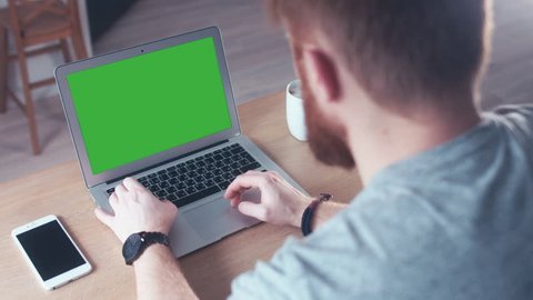 Over the shoulder shot of a young Caucasian man working from home on a laptop, green screen chroma key. 4K UHD 60 FPS slow motion