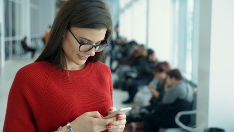 Young woman with smart phone at the airport. She sending sms and looking at the camera. Traveler waiting for the flight