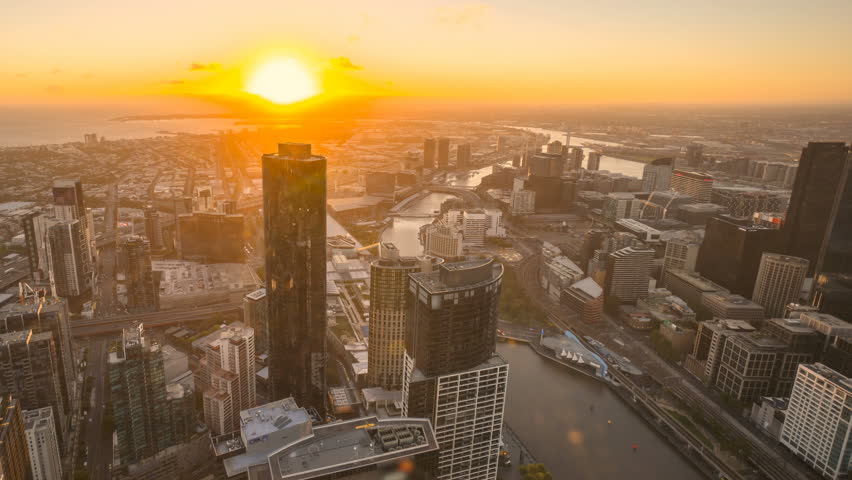 An aerial view of Melbourne cityscape including Yarra River and Victoria Harbour in the distance. Time lapse during sunset with beautiful sun ray bursting through fast moving clouds. Pan right