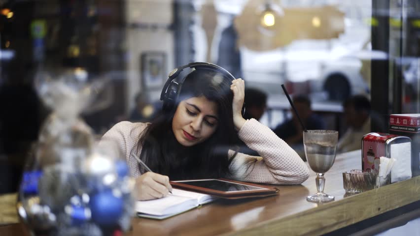 Urban Scene: Middle Eastern girl listening to music in cafe and making notes in notepad. Busy street traffic is on window reflection. Moscow city, Russia. Real time medium shot. Royalty-Free Stock Footage #23346907