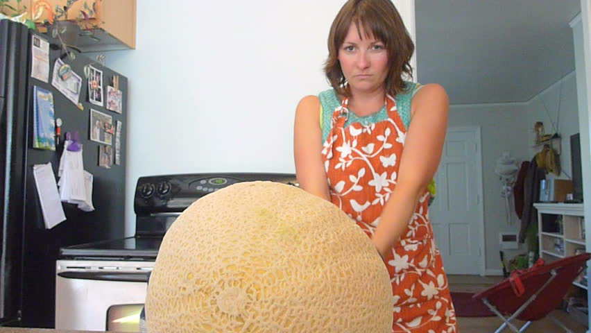 Woman in kitchen with large kitchen knife laughs as she tries to cut cantaloupe