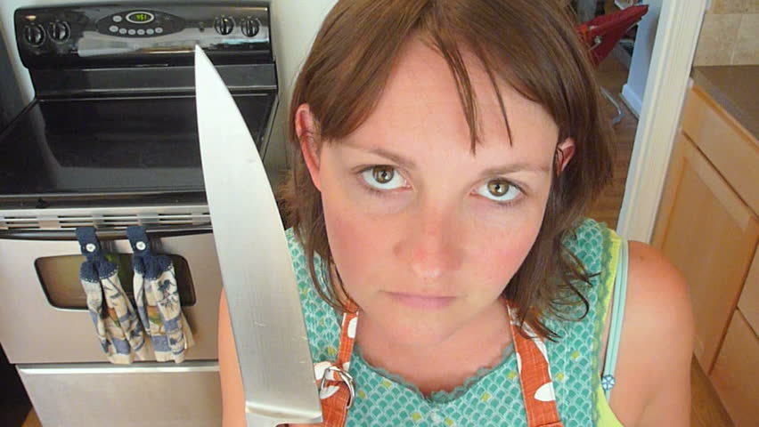 Woman in kitchen with large kitchen knife looks into camera very serious.