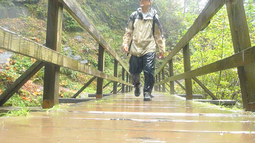 Man in Oregon forest hikes toward camera on wooden bridge during rain storm with