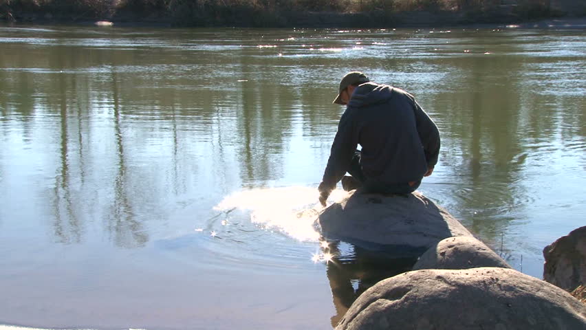 Man on river's edge in Oregon splashing hand reflecting sunshine on water.