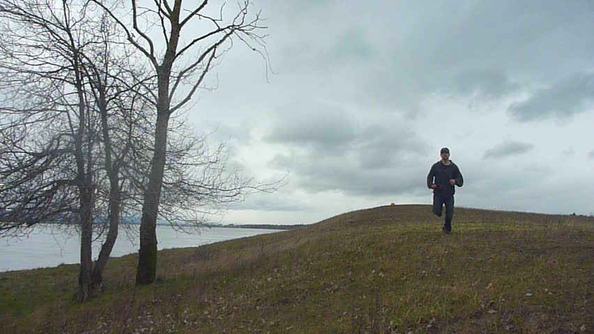 Man running towards camera on hill with tree on a cloudy day by the river.