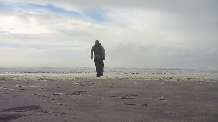 Hiking man walking beach at the Pacific Ocean reaches shore, raises arms and