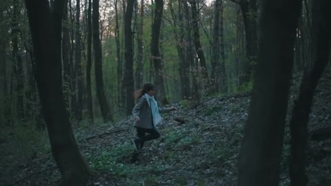 Little European girl with a long hair, brown jacket, black pants, sneakers and bright blue scarf. A scared little child is running through the dark deserted forest. Nightmare, loneliness.