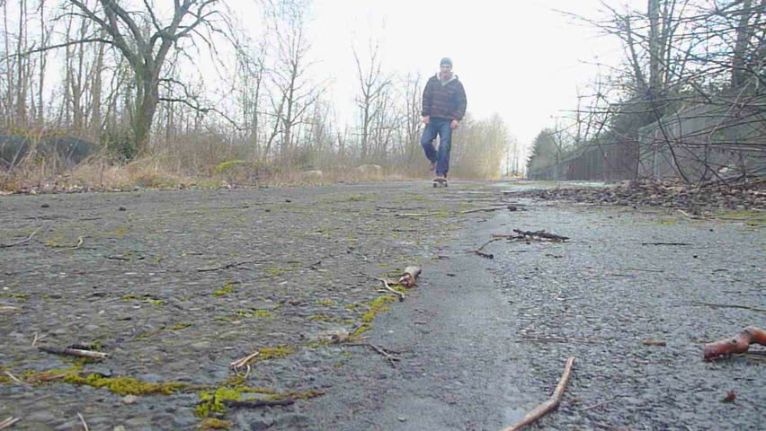 Man skateboards towards and passes camera up old, abandoned road.
