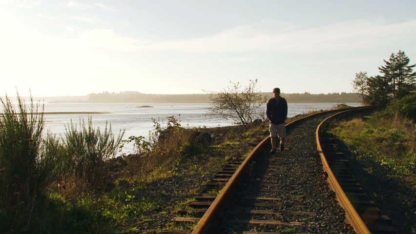 Man walks toward camera along railroad tracks near waterway in Oregon.