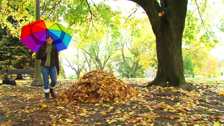 Woman walks by large leaf pile with colorful umbrella during Autumn.