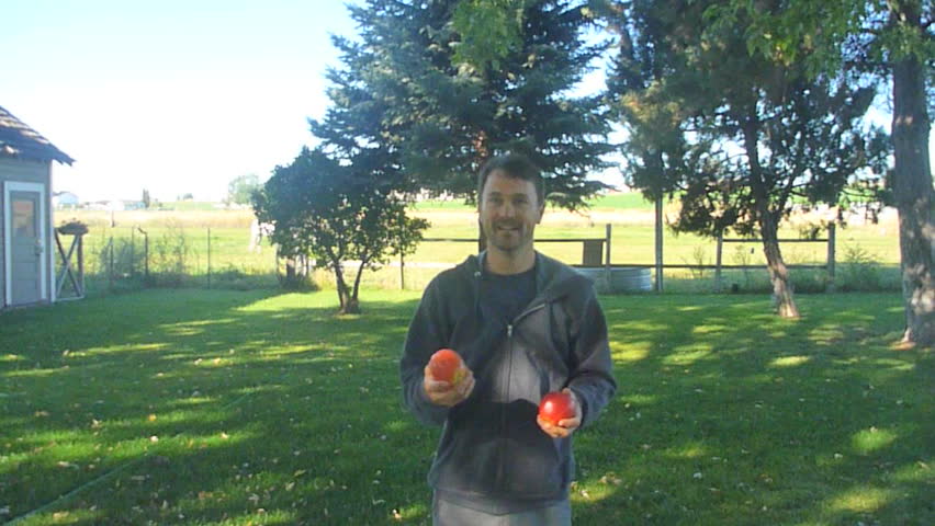 Person outdoors juggles apples and smiles to camera.