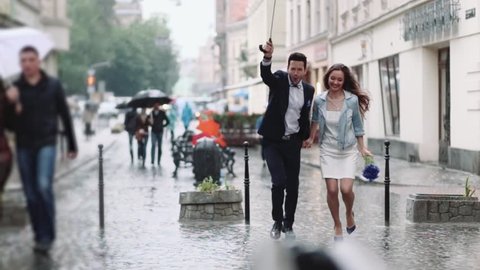 Young handsome bearded man in an elegant costume and tiny attractive woman in casual wear with cornflower bouquet. Young people running under the umbrella in the city center under the heavy rain.