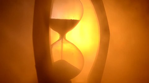 Time concept. Silhouette of Hourglass clock and old vintage wood clock with arrow and smoke on dark background with hot yellow orange  blue cold back lighting, or symbols of time under smoke, the end