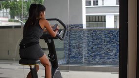 Young Woman Riding Excercise Bike in Gym and Sending Text Message on Phone