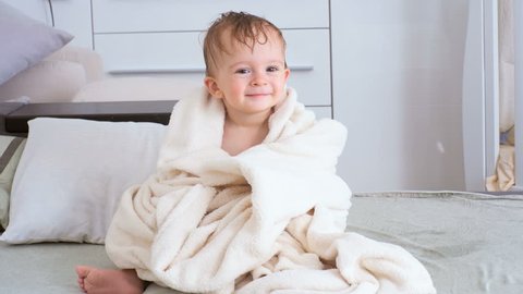 4K footage of cute smiling baby boy covered in towel sitting on bed after bathing