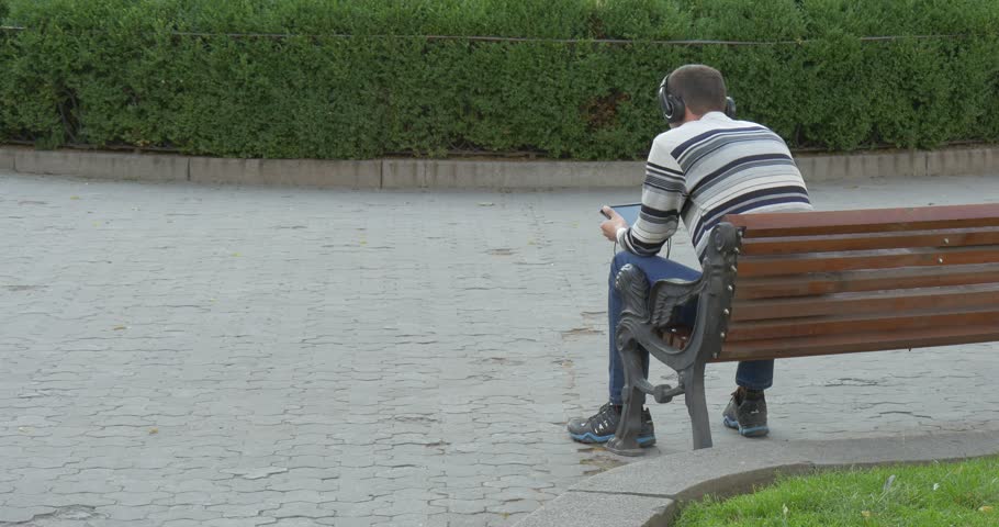 an Adult Man Sits on a Wide Wooden Bench. he is in a Centre of a City Park. the Man Listens to Music and Reads a Book.