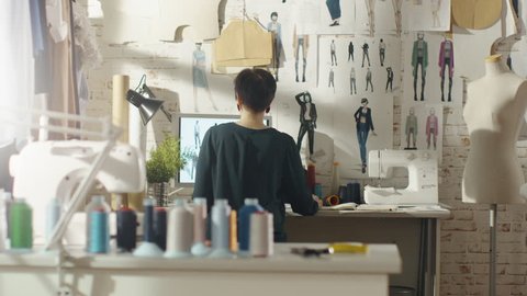 Backview of a Female Fashion Designer Working on a Personal Computer, Taking Notes. Her Workplace is Sunny, Sewing Machine Standing on Her Table, Sketches of Future Collection Pinned to the Wall.
