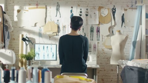 Zoom Out of a Female Fashion Designer Looking at Drawings and Sketches that are Pinned to the Wall Behind Her Desk. Studio is Sunny. Personal Computer, Colorful Fabrics, Sewing Items are Visible.