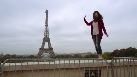 Paris, France - October 15, 2016: Smiling girl stand on concrete fence and pose for banal touristic photography with Eiffel Tower on cloudy day. Slow motion. Travel activities concept.