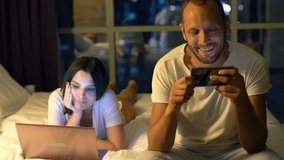 Young couple watching movies on smartphone and laptop in bedroom at night
