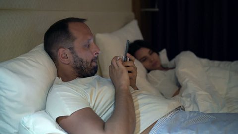 Couple in bed at night, man cheating, texting on smartphone while woman sleeping

