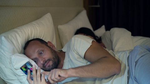 Young couple fighting, arguing in bed at night, man texting on smartphone 
