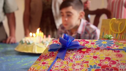 Group of happy children celebrating birthday at home, kids and friends having fun at party. Child blowing candles on cake, table full of boxes, gifts, presents. Slow motion
