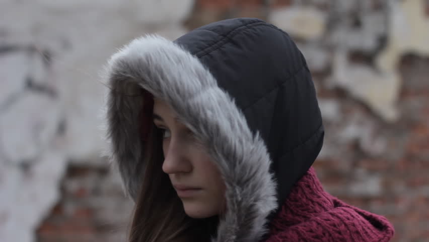 desperate teen: Close up of sad and lonely girl with hood up against old brick wall of abandoned building in cold dark weather, steadicam shot