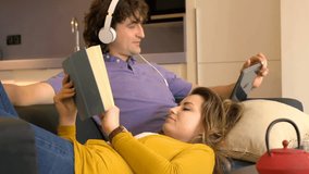 Woman reading book on her boyfriend's knees who is watching video

