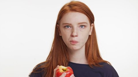 Concentrated red-haired teenage Caucasian girl eating apple chewing fast on white background