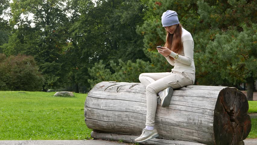 Young adult woman sit resting alone in park, stare to smartphone. Hipster girl ensconce lop down on large log, texting on mobile phone. Green lawn and bushes on background. Relaxed lady dangle on feet | Shutterstock HD Video #23479714