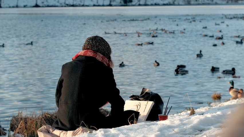 People in the city park at winter season. Young artist painter woman sits on snow lake shore and paints a picture with paints and brush. On the background of water with many birds ducks