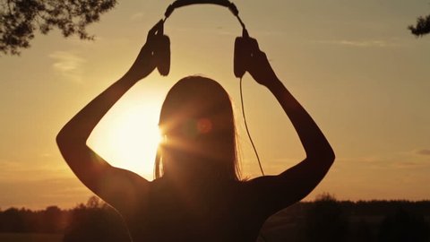 Silhouette of woman putting on the headphones, listening to music and dancing in the forest. Sunset light, sun lens flares, golden hour. Relax, nature and joyful concept