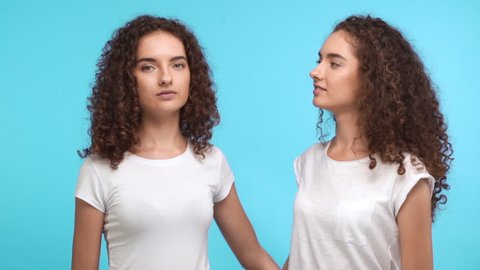 Two charming Caucasian curly female twins in white t-shirts embracing each other on shoulders smiling and showing ok with thumbs on blue background in slowmotion