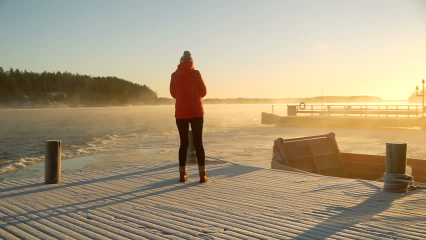 young travel woman go down and stay on wooden dock covered with ice on frozen lake near the shore and wave water during strong snowstorm and high wind in cold winter day