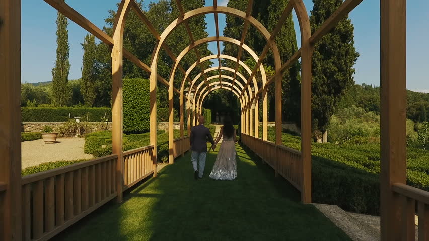 Man in grey suit and woman in long dress walk under archs for grape in the garden