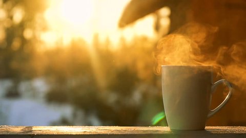 female young hand put white mug of hot drink with steam on wooden desk of stoop of house in the country near forest during winter snowy day under sunlight of sunset