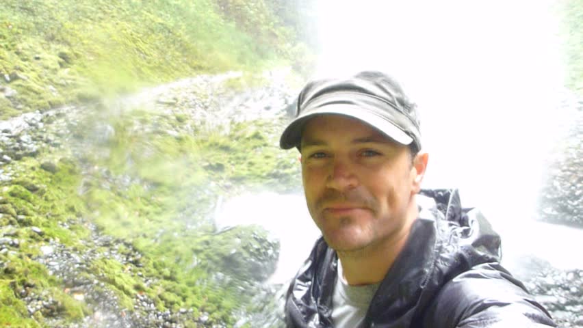Man under large waterfall in Oregon on rainy day waives hand to camera and looks