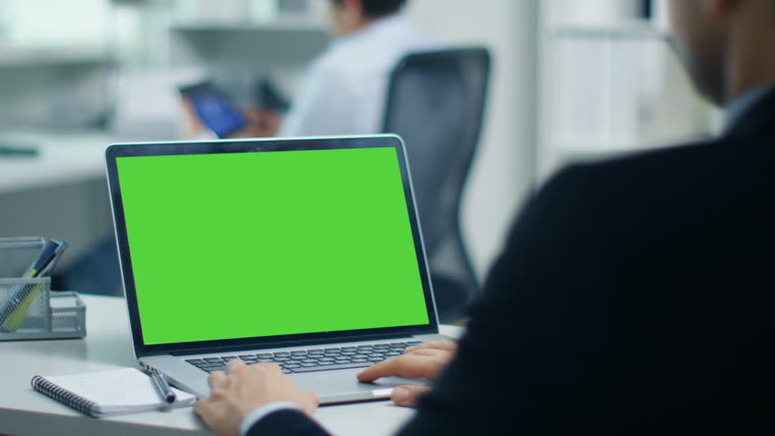 Businessman Working on a Laptop with Green Screen on. In the Background His Colleague Working on a Tablet Computer. Office is Modern and Bright. Shot on RED Cinema Camera 4K (UHD).